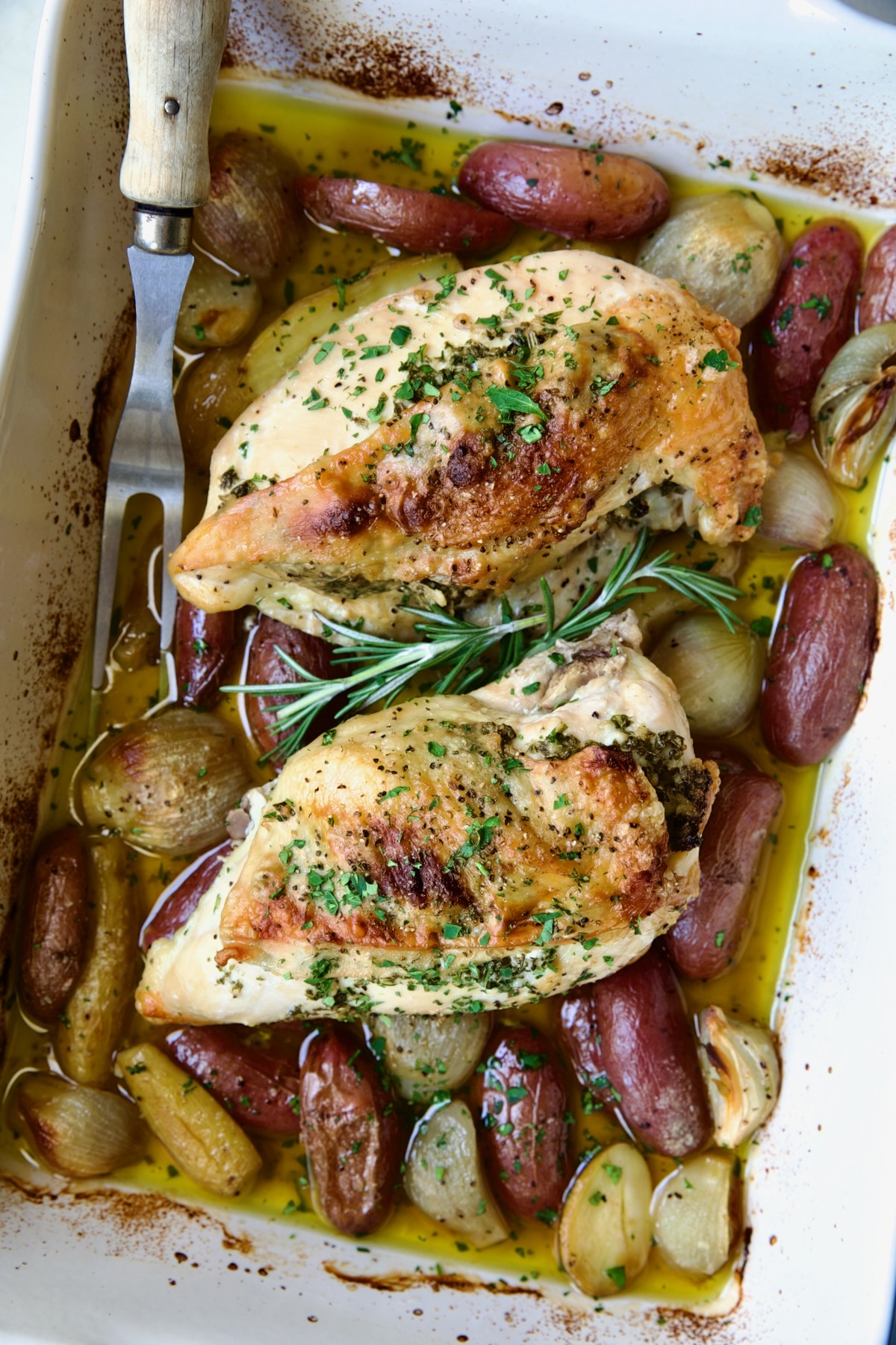 Two Roast Split Chicken Breasts with Potatoes and Shallots in white rectangular baking dish.