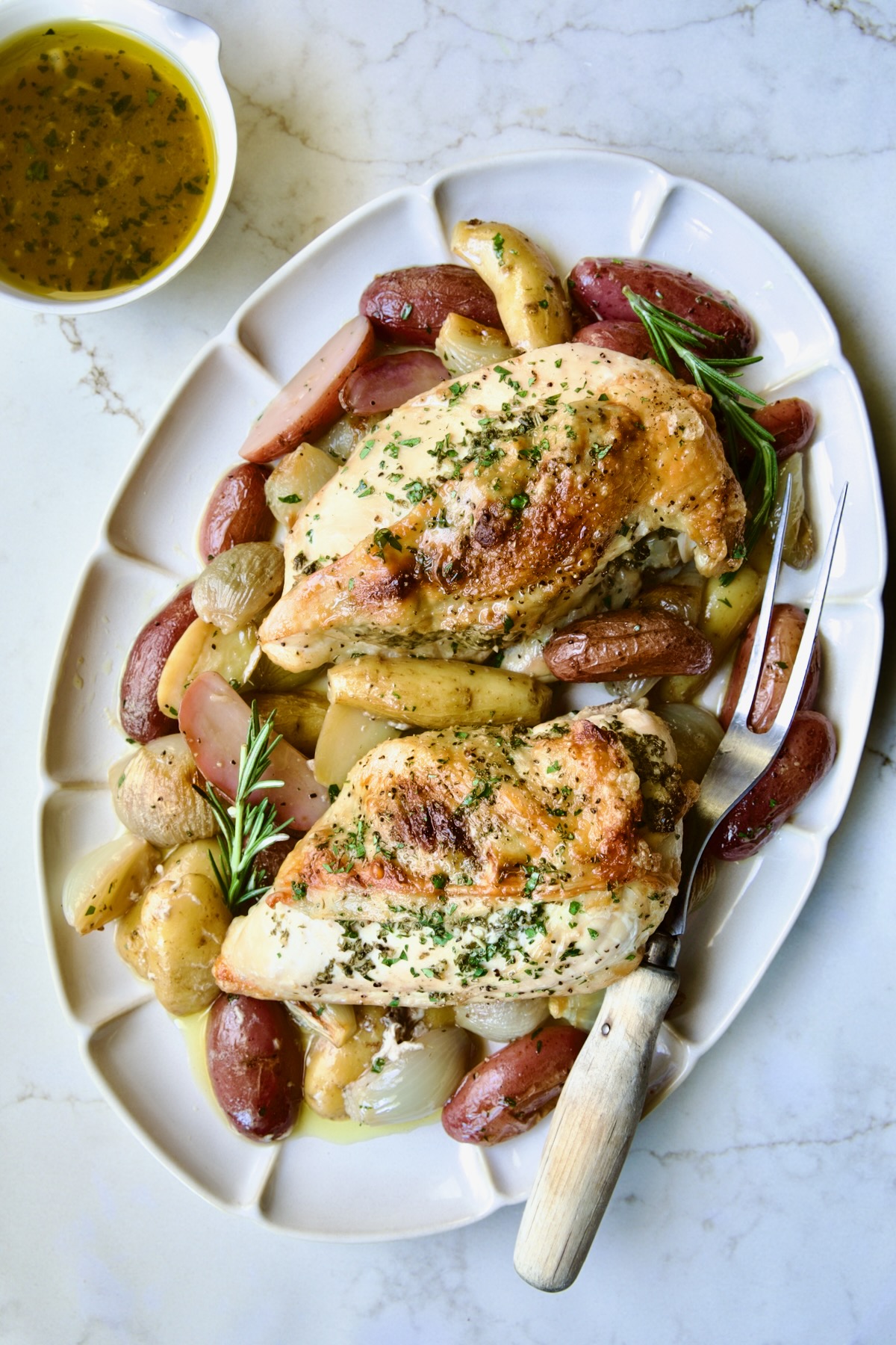 Two Roast Split Chicken Breasts with Potatoes and Shallots on white oval platter with meat fork.