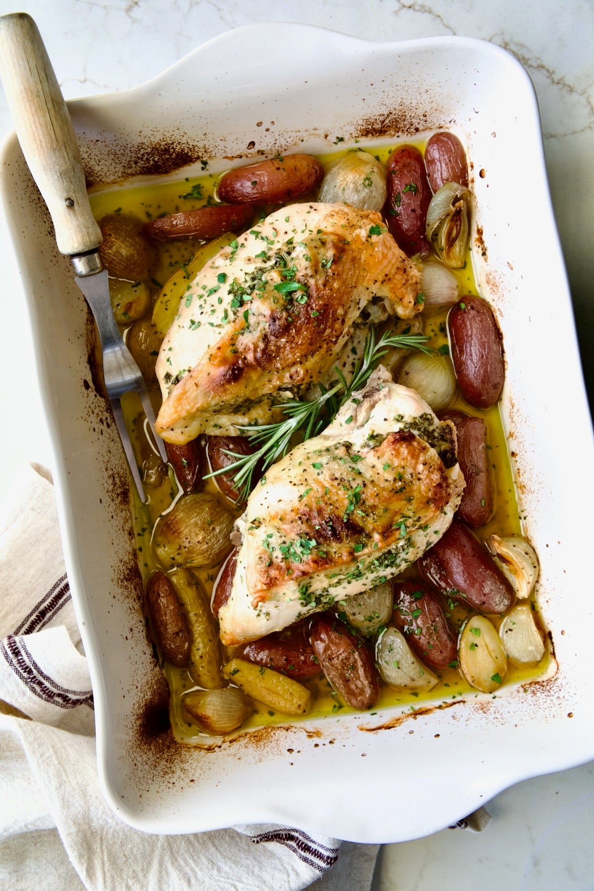 Two Roast Split Chicken Breasts with Potatoes and Shallots in white rectangular baking dish.