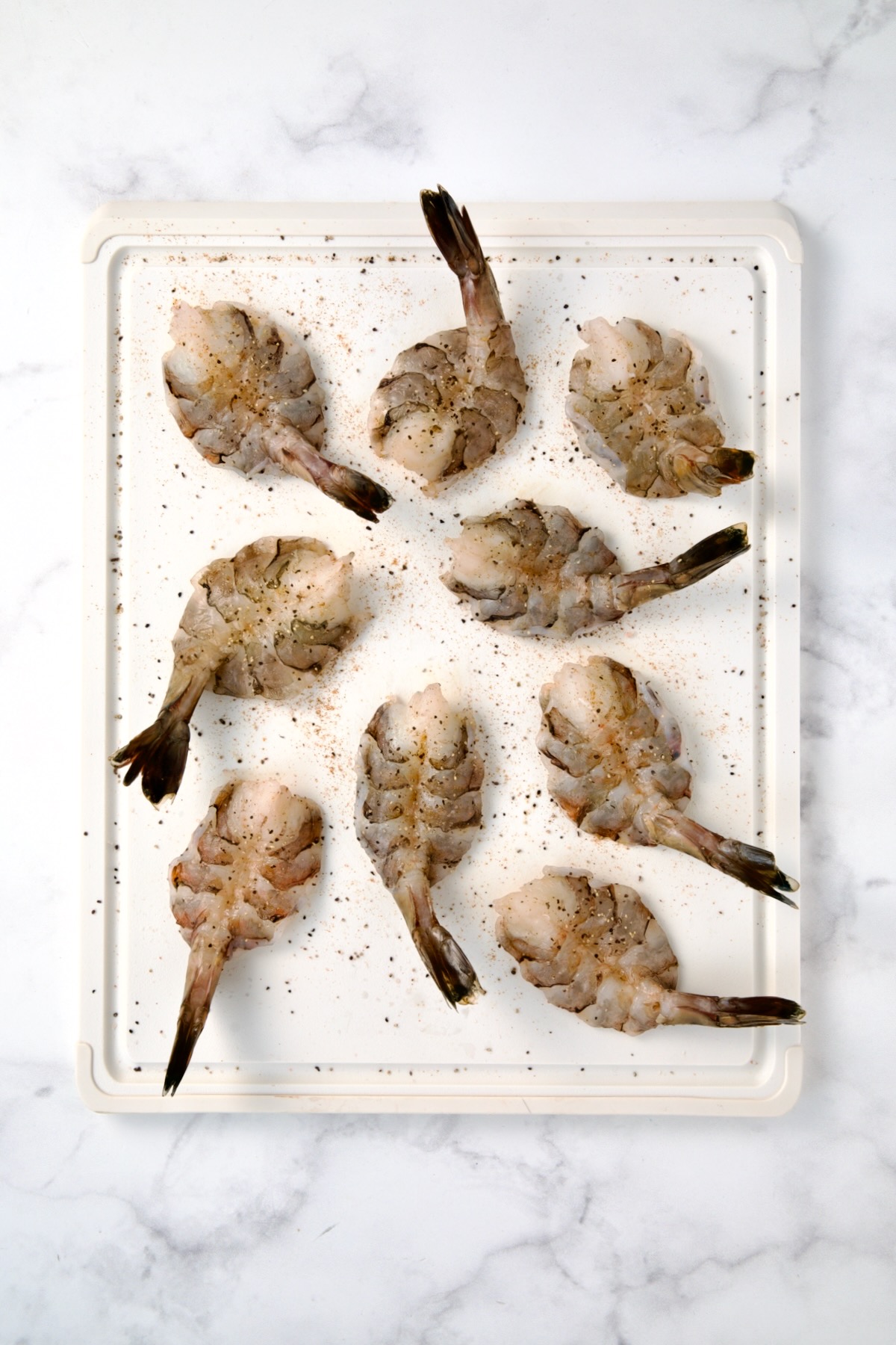 Nine colossal peeled and deveined shrimp on white plastic cutting board after being butterflied seasoned with salt and pepper.