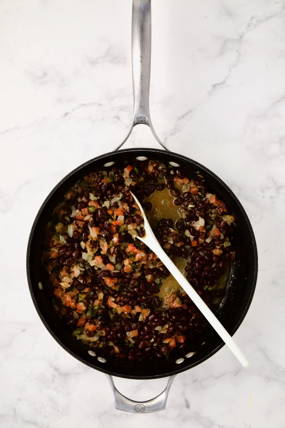 Chicken broth and black beans added to vegetables in nonstick skillet being stirred with white spoon.