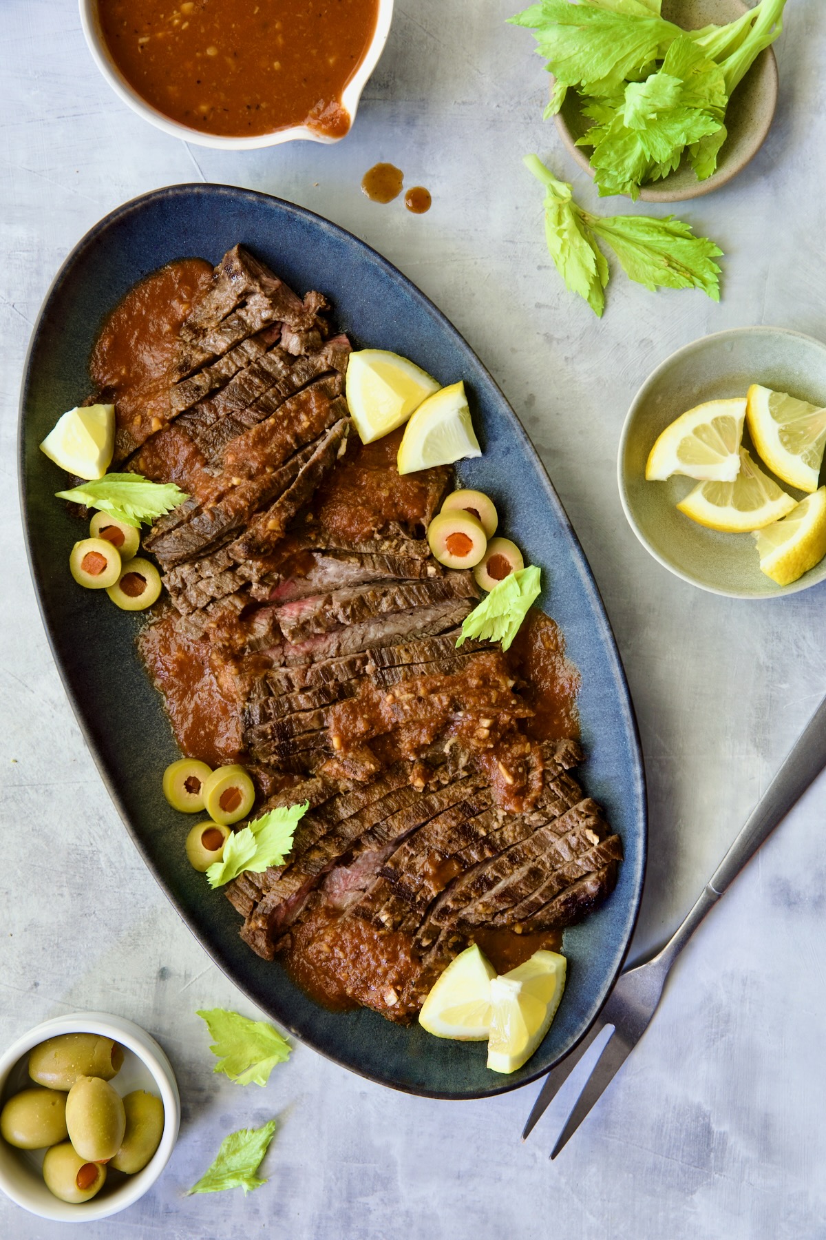 Sliced Bloody Mary Skirt Steak on blue oval platter garnished with lemon wedges, olive slices and celery leaves.