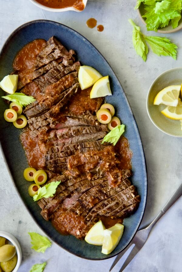 Sliced Bloody Mary Skirt Steak on blue oval platter garnished with lemon wedges, olive slices and celery leaves.