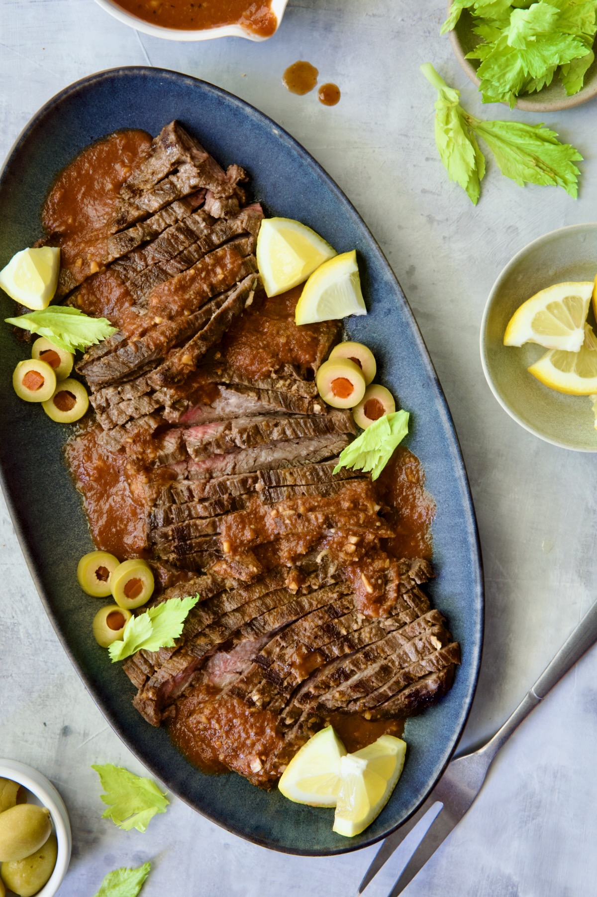 Sliced Bloody Mary Skirt Steak on blue oval platter garnished with lemon wedges, olive slices and celery leaves.