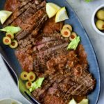 Sliced Bloody Mary Skirt Steak on blue oval platter garnished with lemon wedges, olive slices and celery leaves.