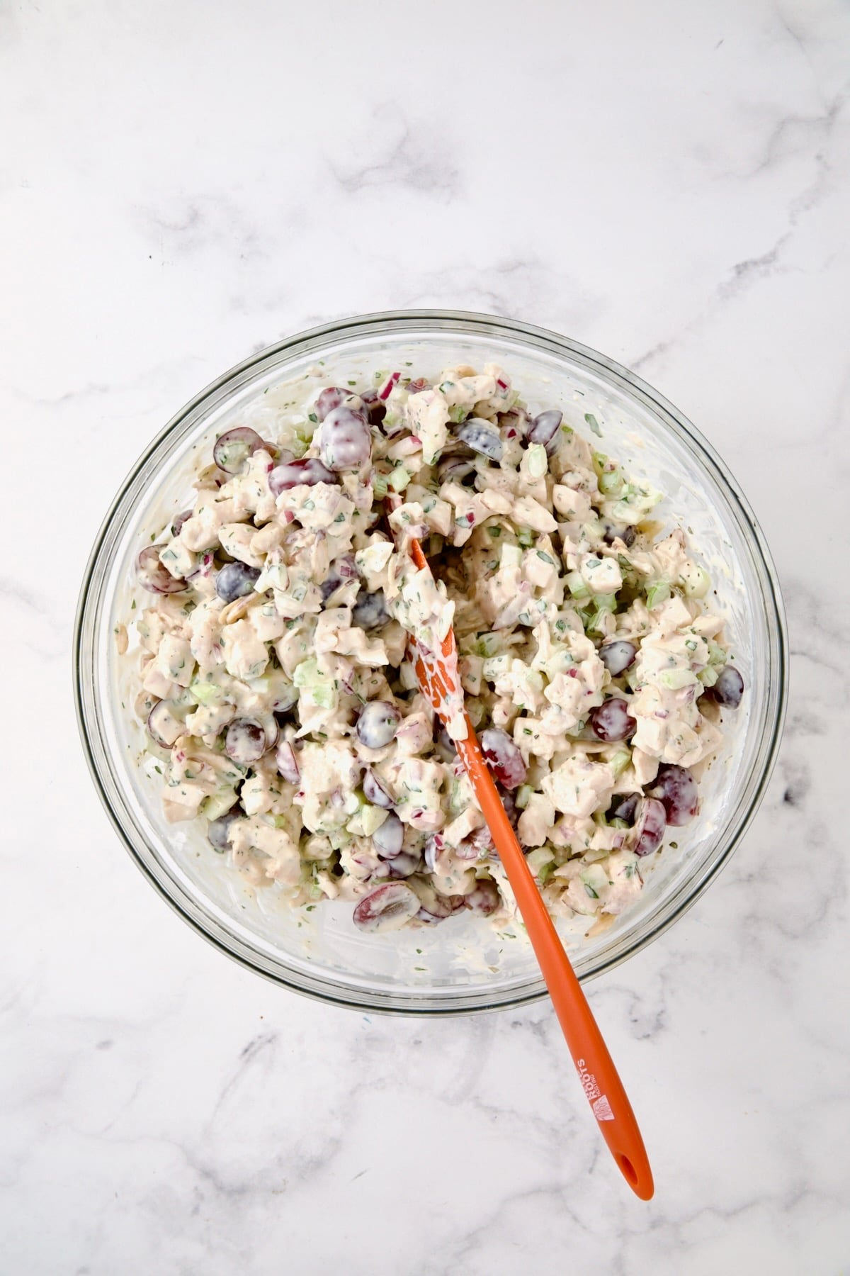 Chicken salad in glass bowl being stirred with orange spatula.