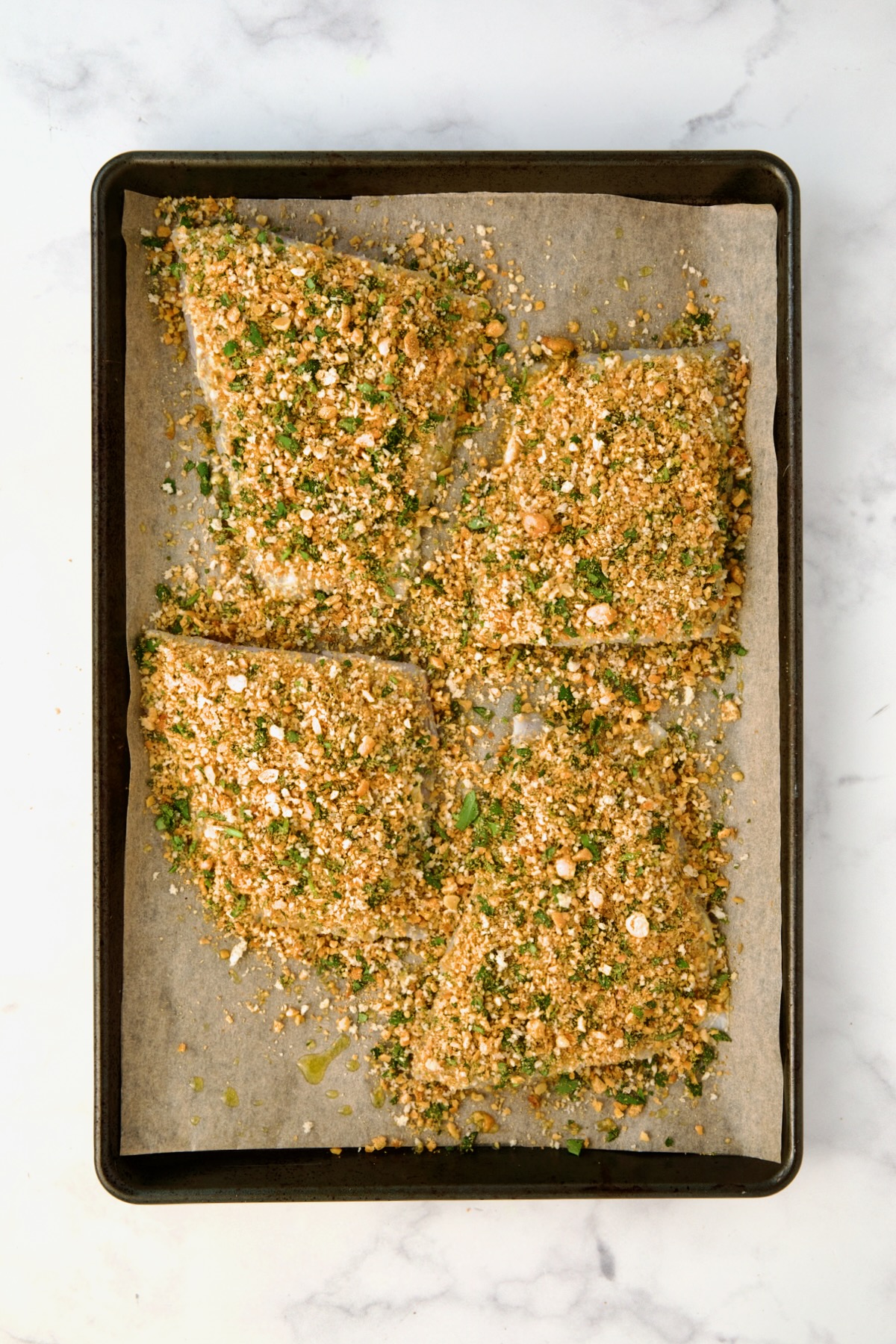 Four pieces of fish on parchment paper-lined baking sheet topped with chickpea topping before being baked.