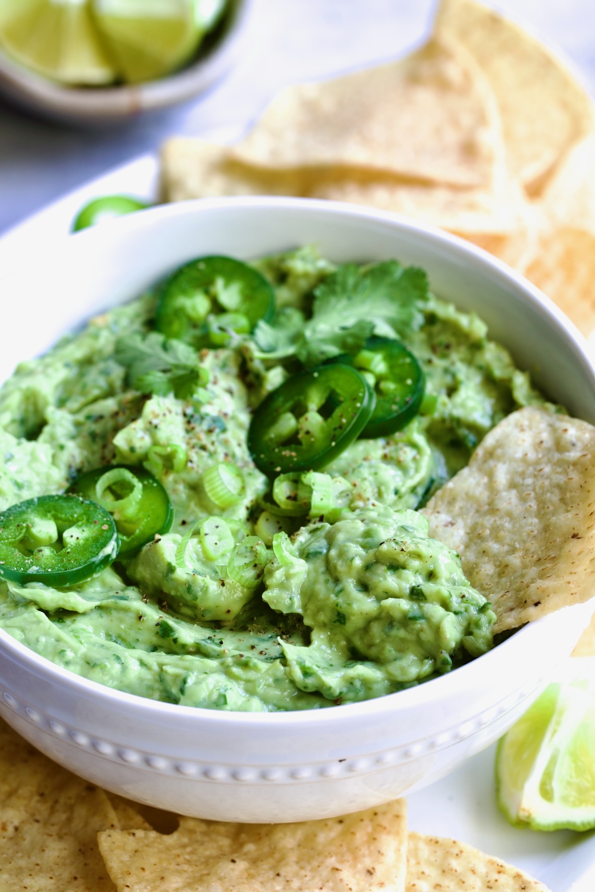 Spicy Guacamole in white bowl surrounded by tortilla chips garnished with jalapeno slices and cilantro leaves.