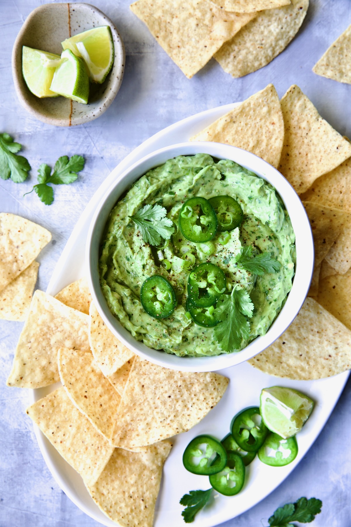 Spicy Guacamole in white bowl surrounded by tortilla chips garnished with jalapeno slices and cilantro leaves.