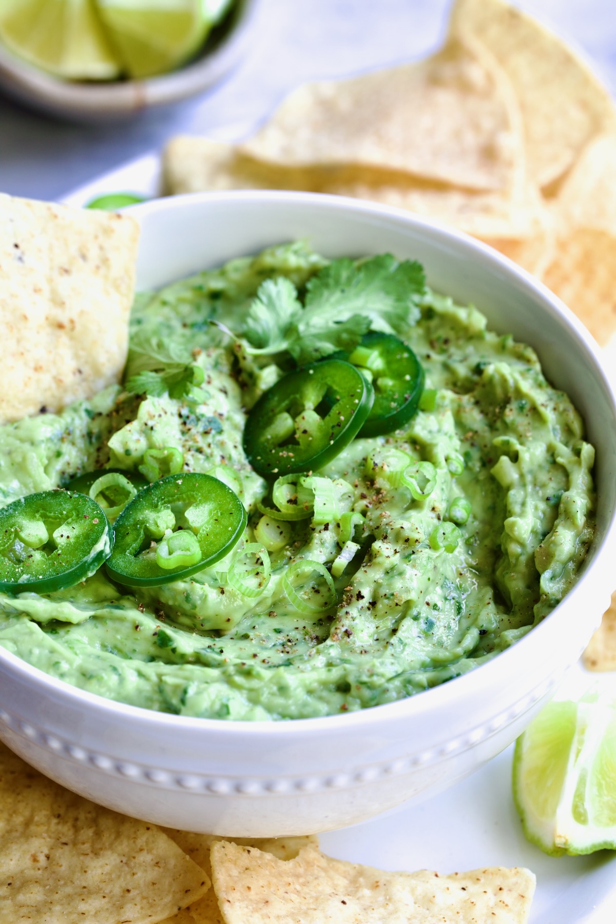 Spicy Guacamole in white bowl surrounded by tortilla chips garnished with jalapeno slices and cilantro leaves.