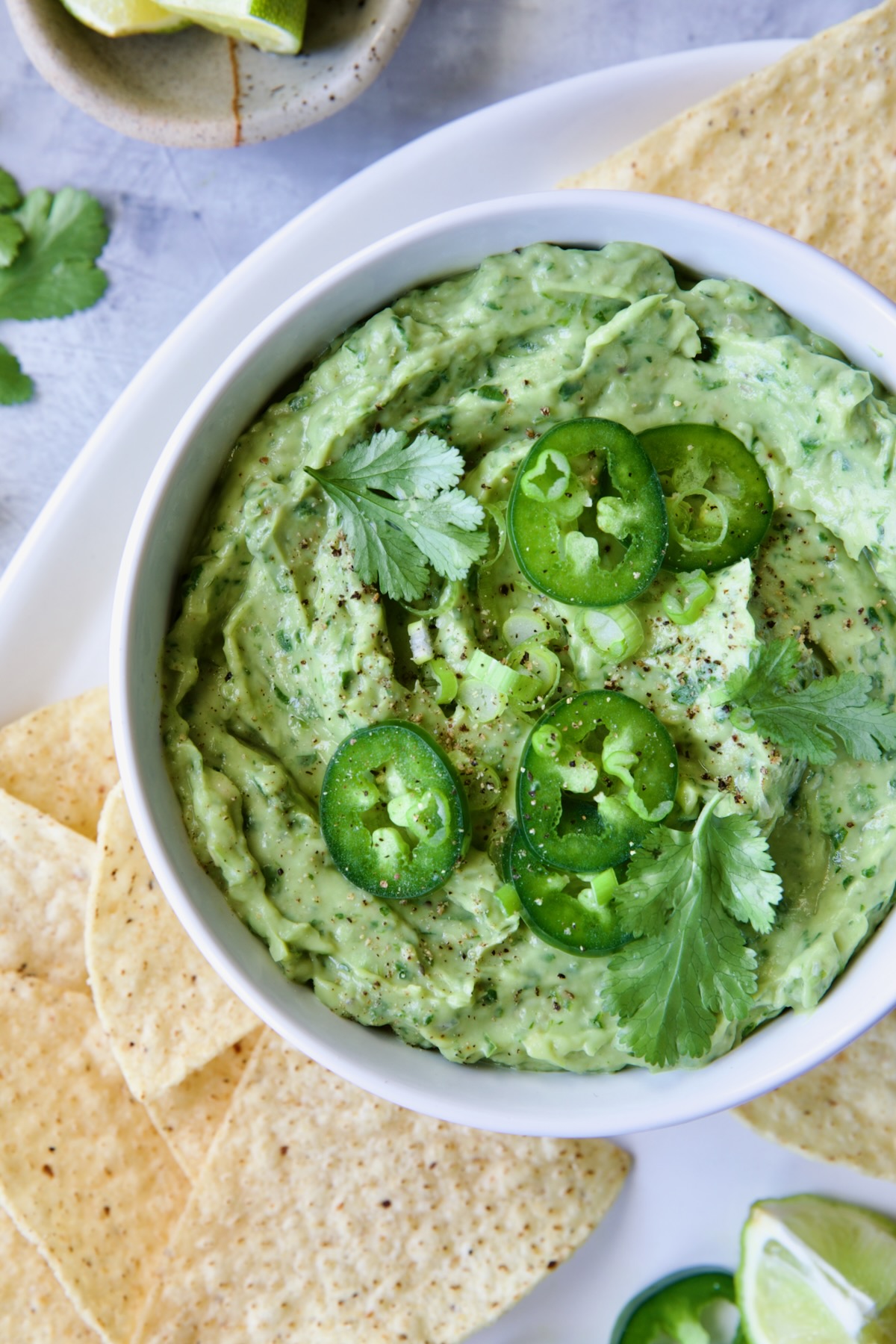 Spicy Guacamole in white bowl surrounded by tortilla chips garnished with jalapeno slices and cilantro leaves.