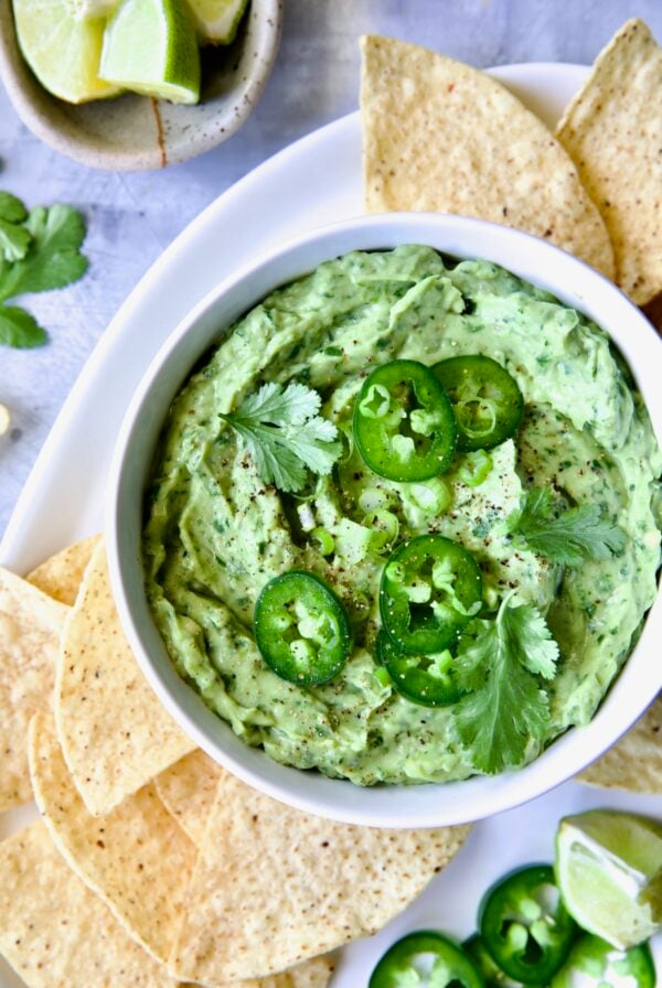 Spicy Guacamole in white bowl surrounded by tortilla chips garnished with jalapeno slices and cilantro leaves.