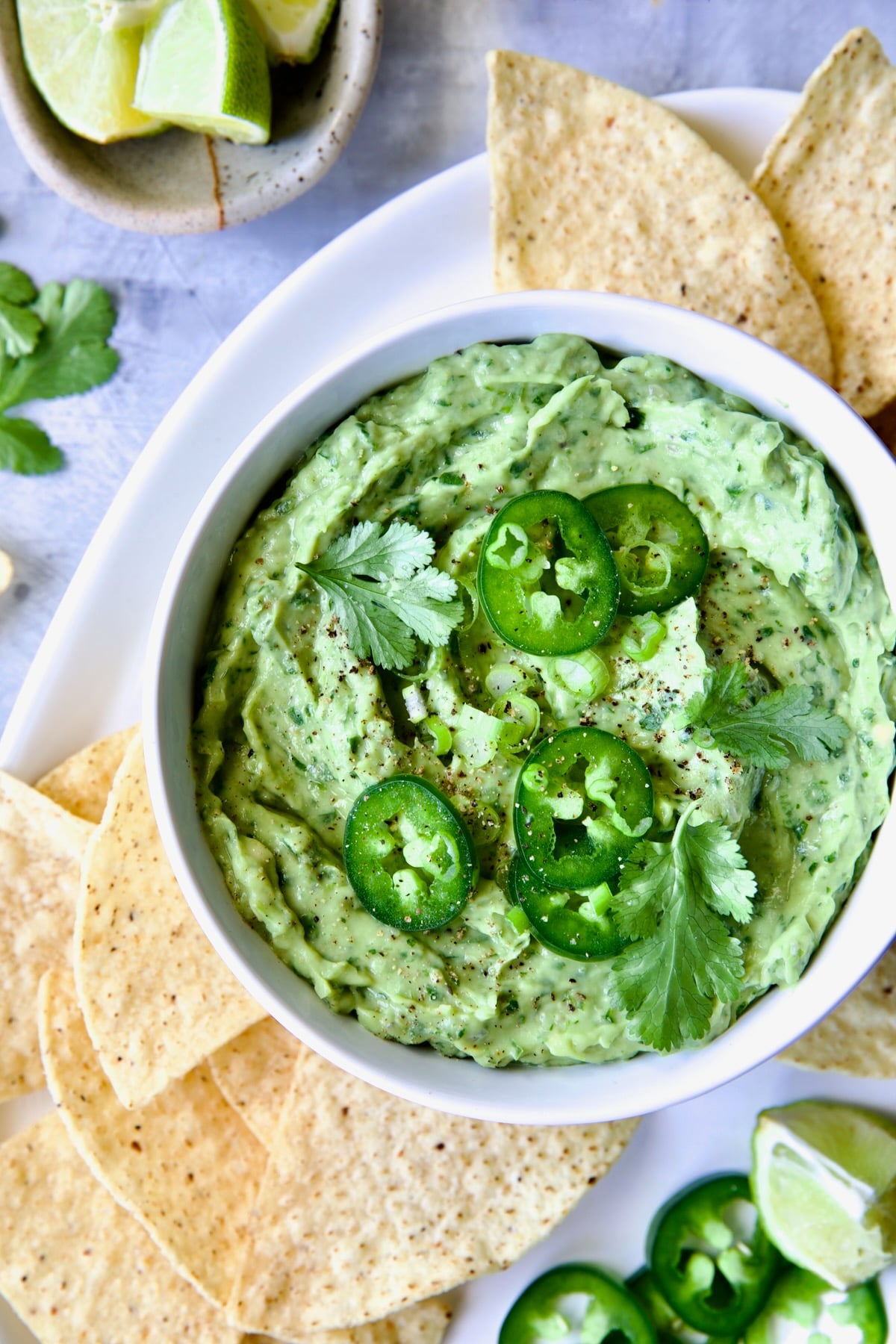 Spicy Guacamole in white bowl surrounded by tortilla chips garnished with jalapeno slices and cilantro leaves.