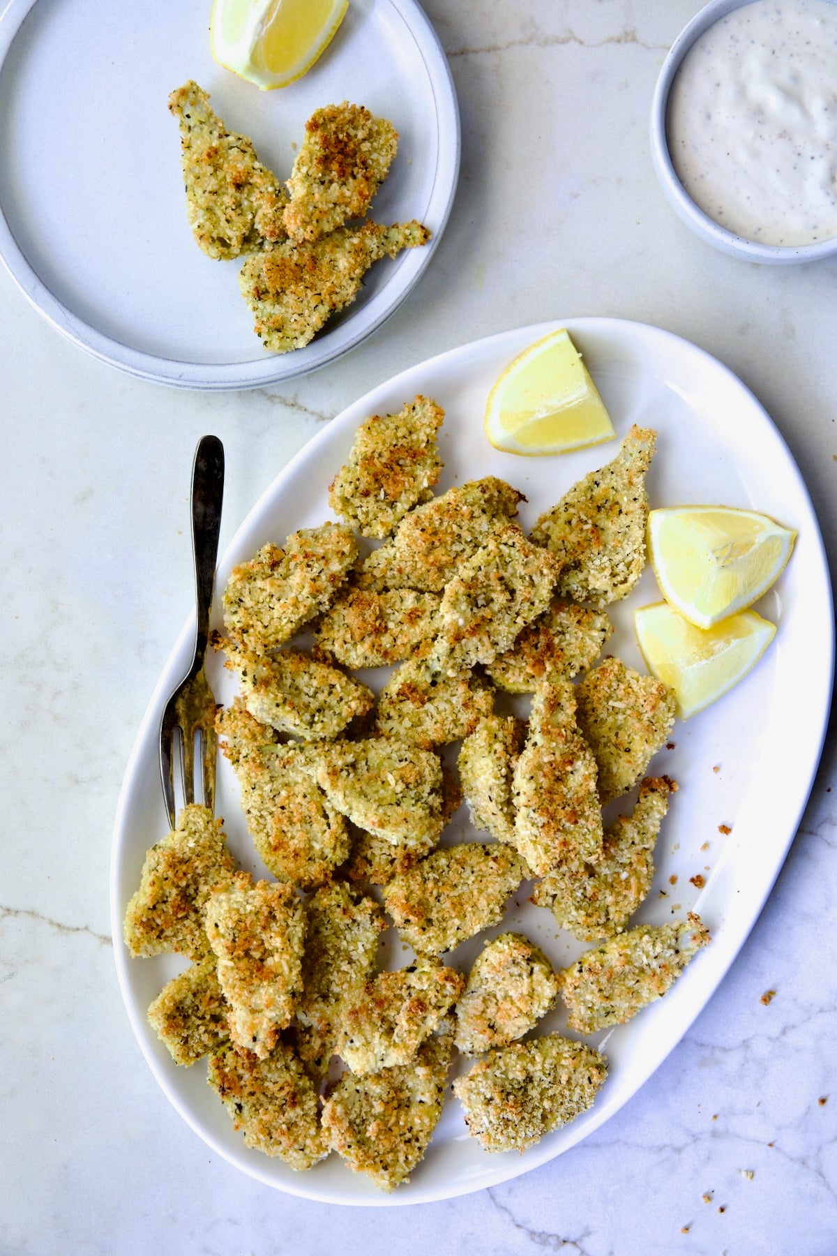 Crispy Parmesan Artichoke Hearts on oval white platter wiht some placed on a small plate with lemon wedges.