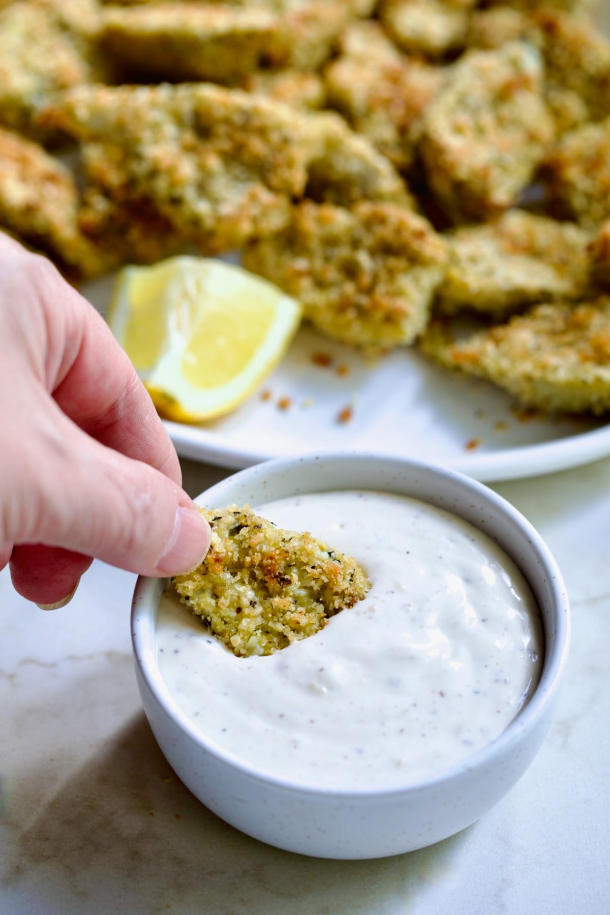 Hand dipping an artichoke heart into the Caesar dip in a white bowl.