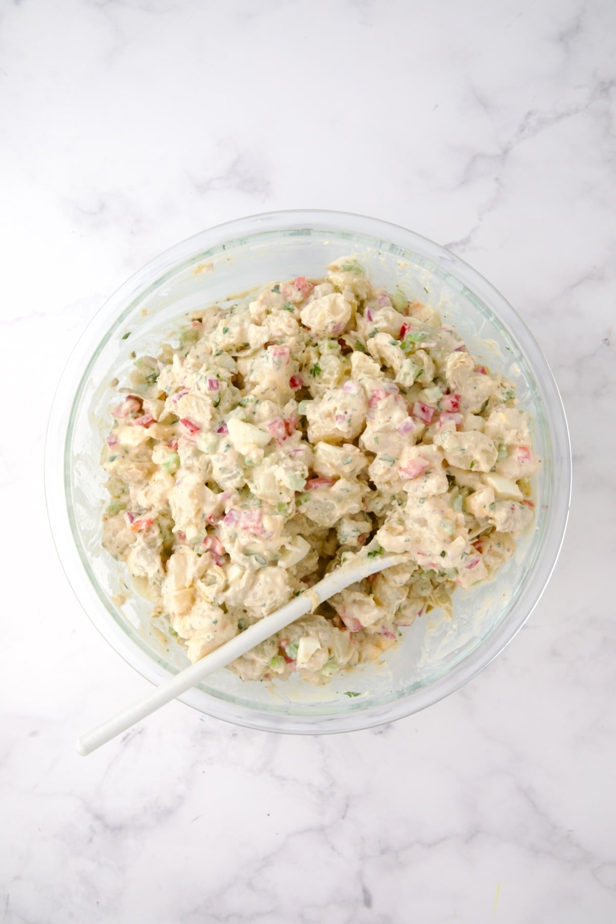 Potato salad in glass bowl with white mixing spoon.