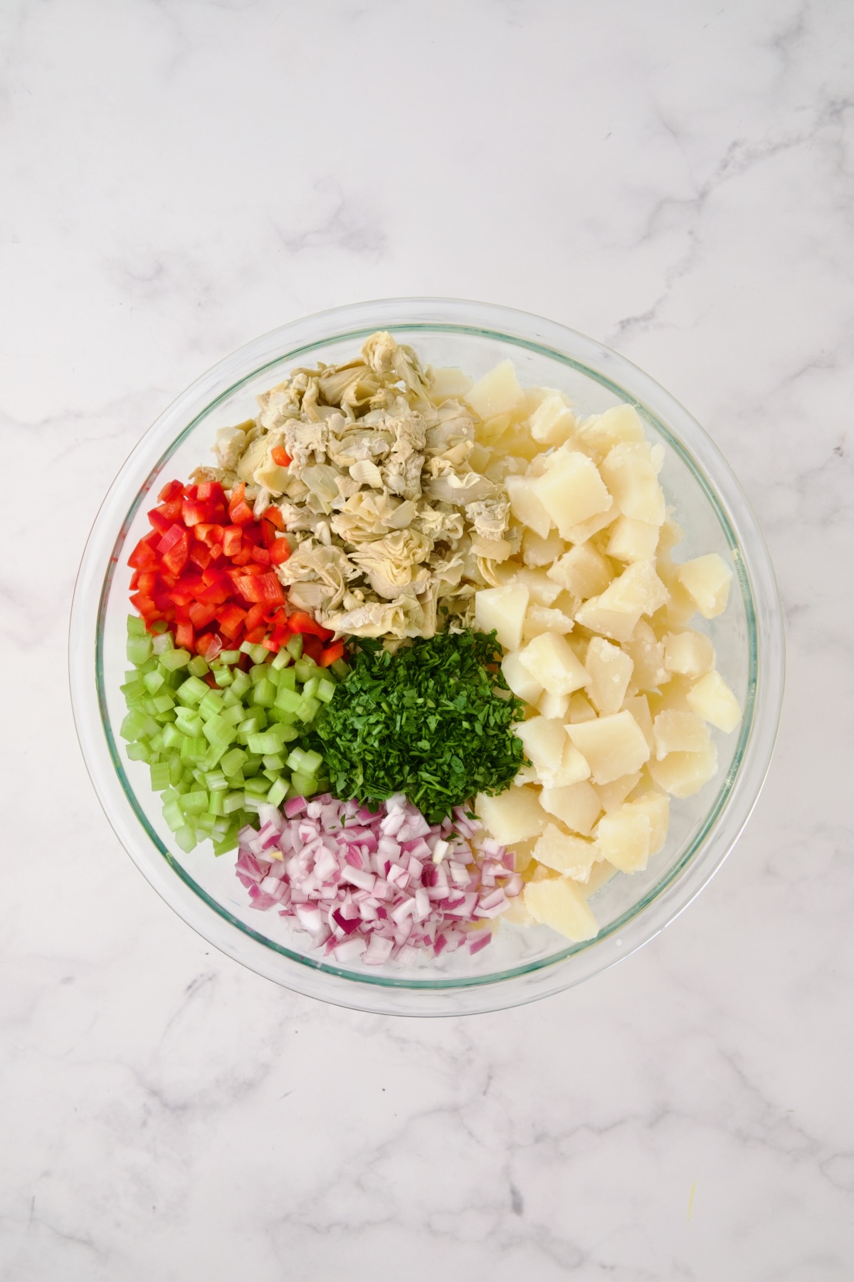 Potatoes, red onion, celery, red bell pepper, artichokes and parsley in glass bowl before being mixed together.