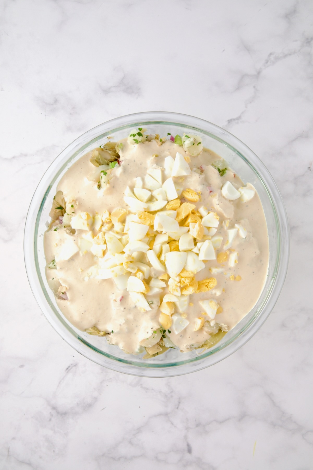 Potatoes, red onion, celery, red bell pepper, artichokes, and parsley in a glass bowl after being mixed together with dressing and eggs added.