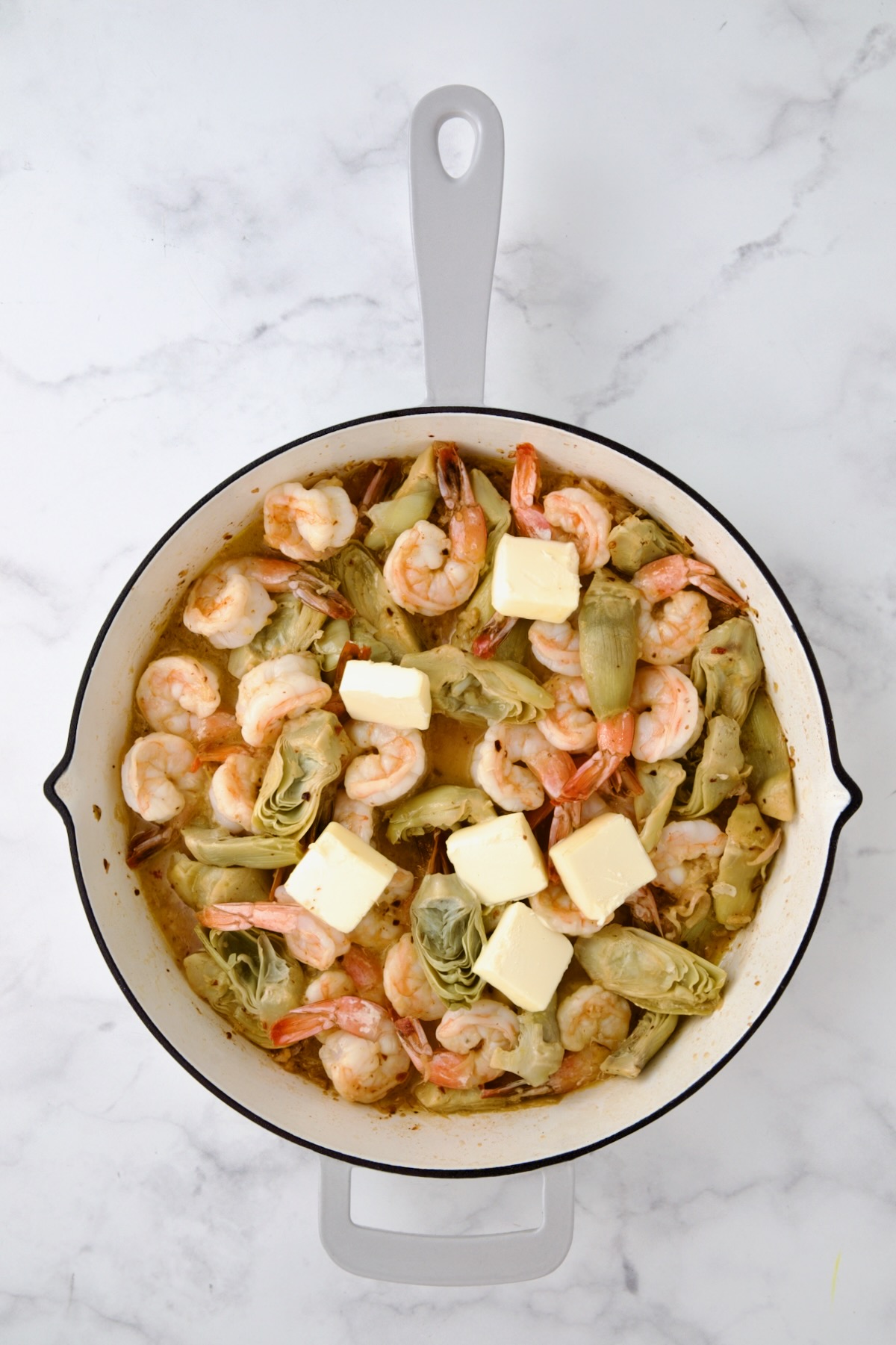 Butter added cooked shrimp and artichokes in white enamel cast iron pan.