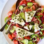 Classic Greek Salad with broken chunks of feta triangles on white oval platter with serving utensils.