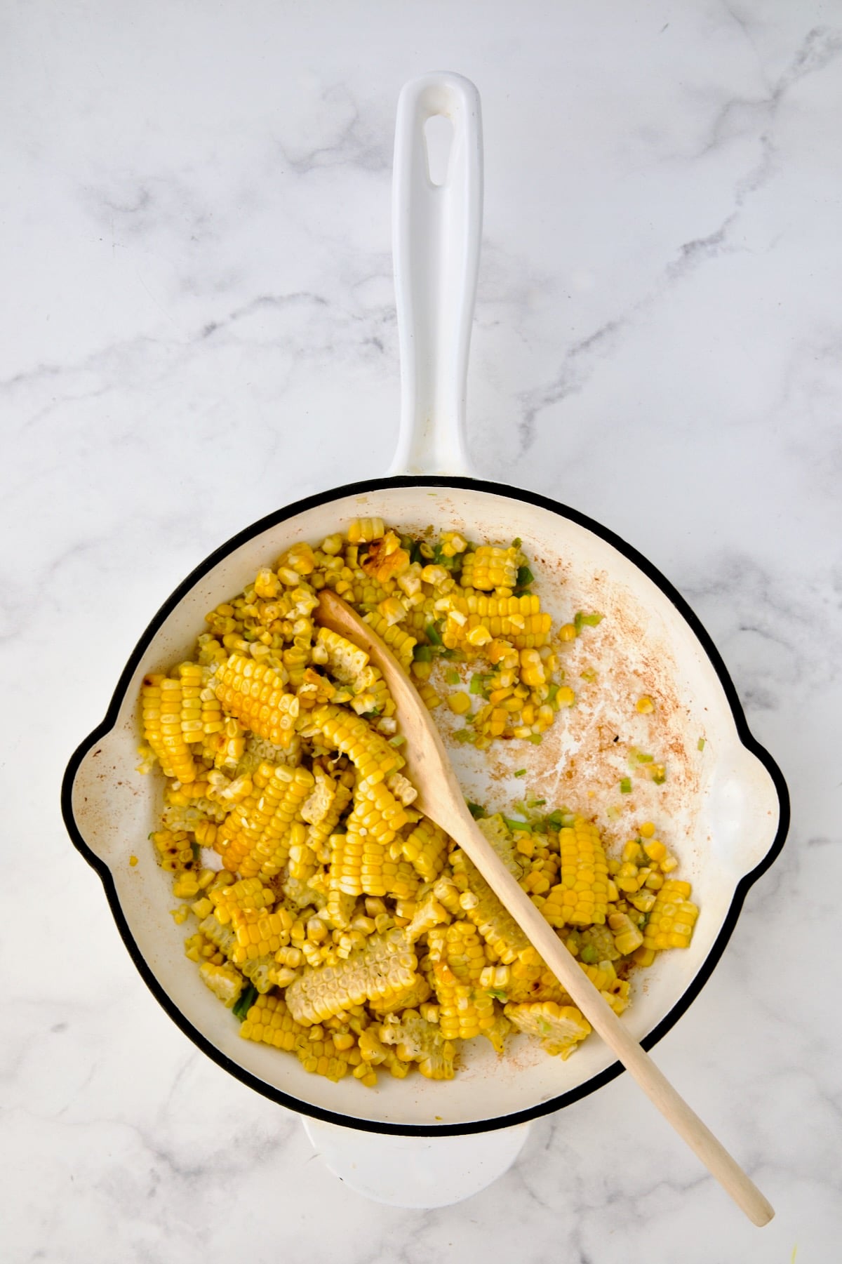 Corn and scallions being stirred with mixing spoon in white enamel cast iron skillet.
