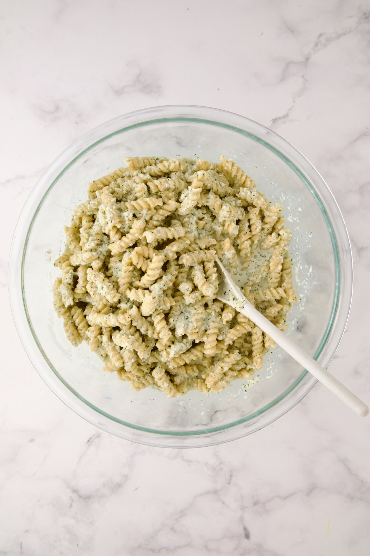 Pasta salad with dressing being mixed together in glass bowl with white mixing spoon.