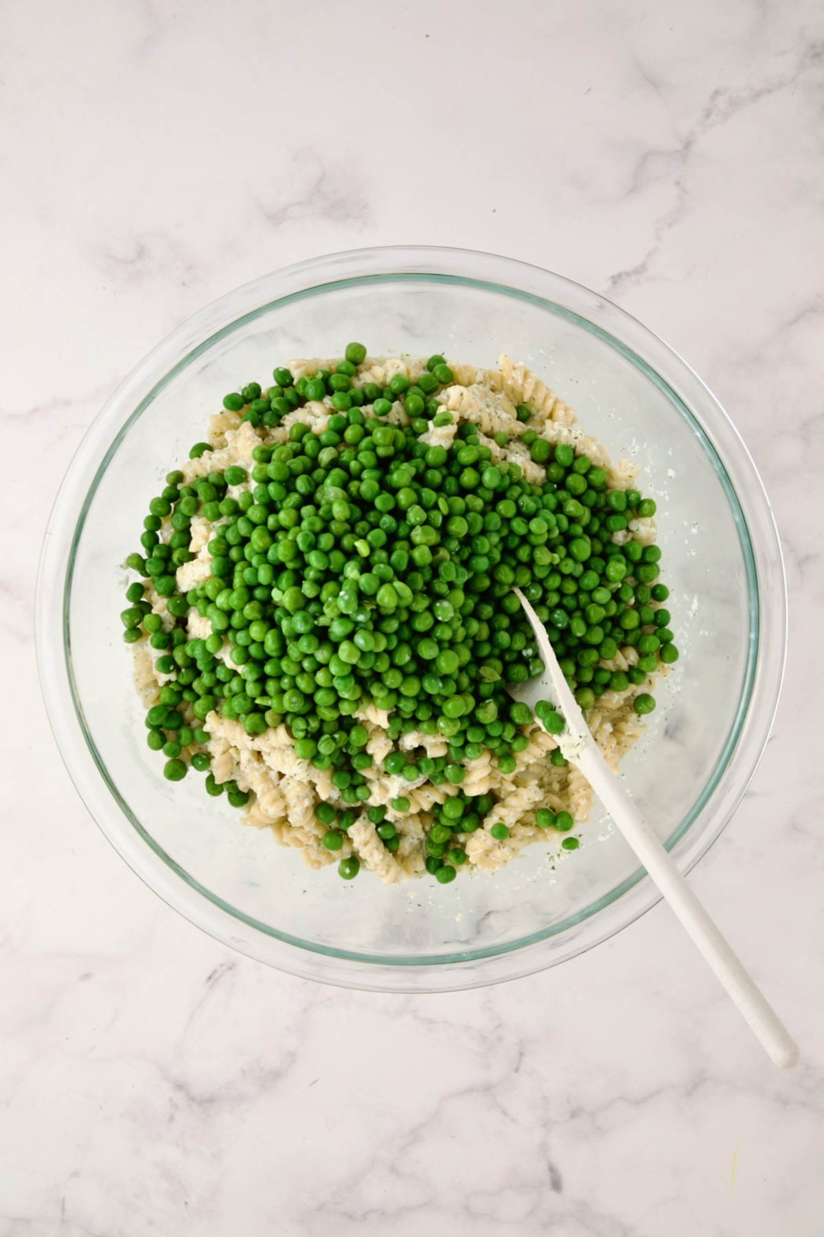 Peas added to pasta salad in glass bowl.