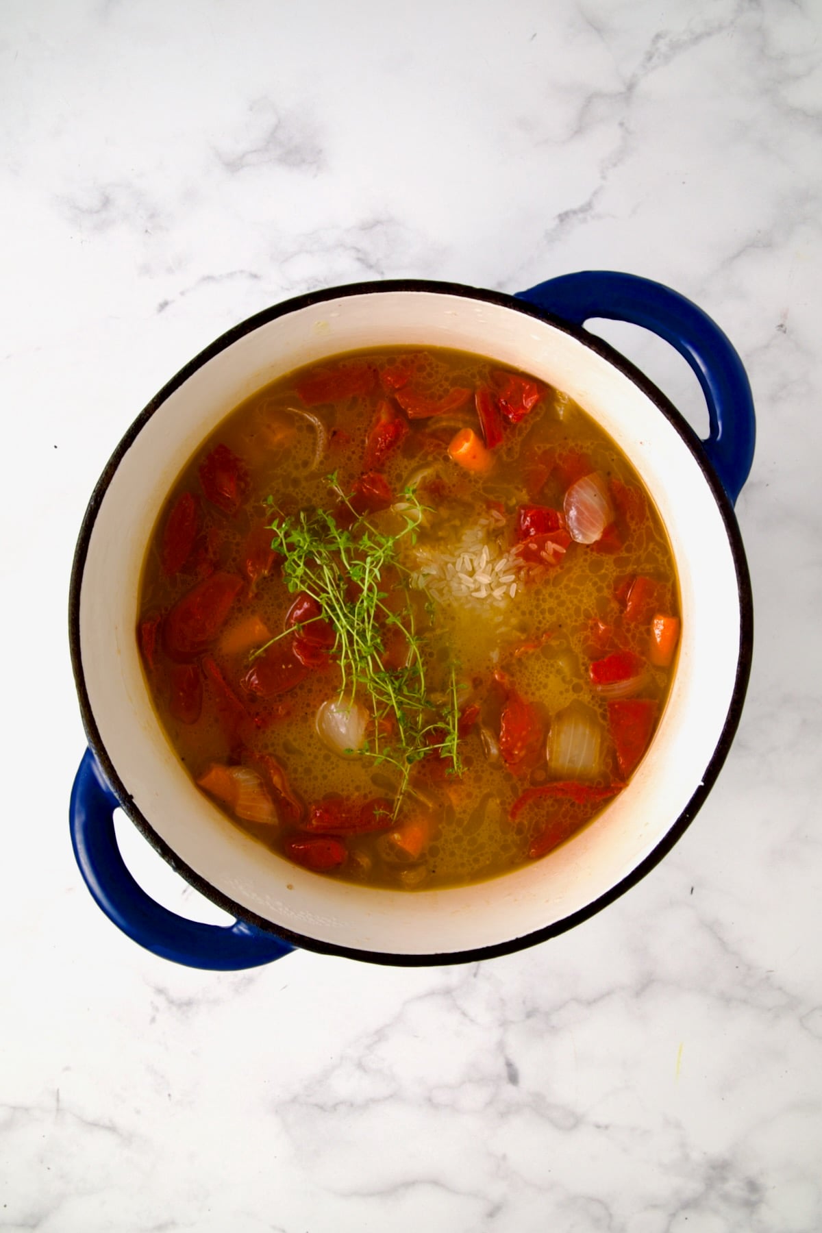 Chunks of tomatoes, onion, carrots and garlic in blue Dutch oven with broth, fresh thyme and rice added.
