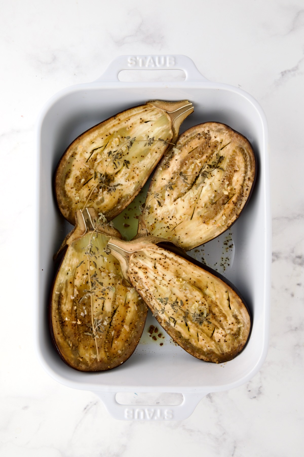 Four eggplant halves in white rectangular baking dish with fresh garlic and herbs over each half after being baked.