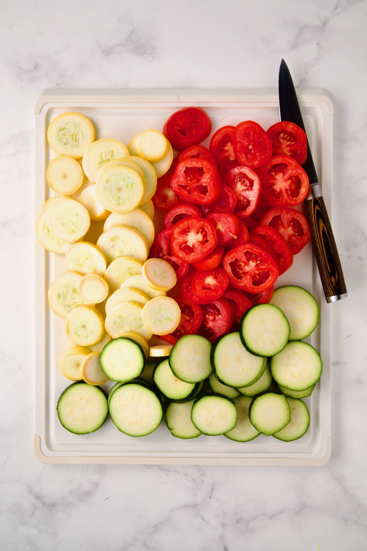 Sliced yellow squash, zucchini and tomatoes on white plastic cutting board with knife.