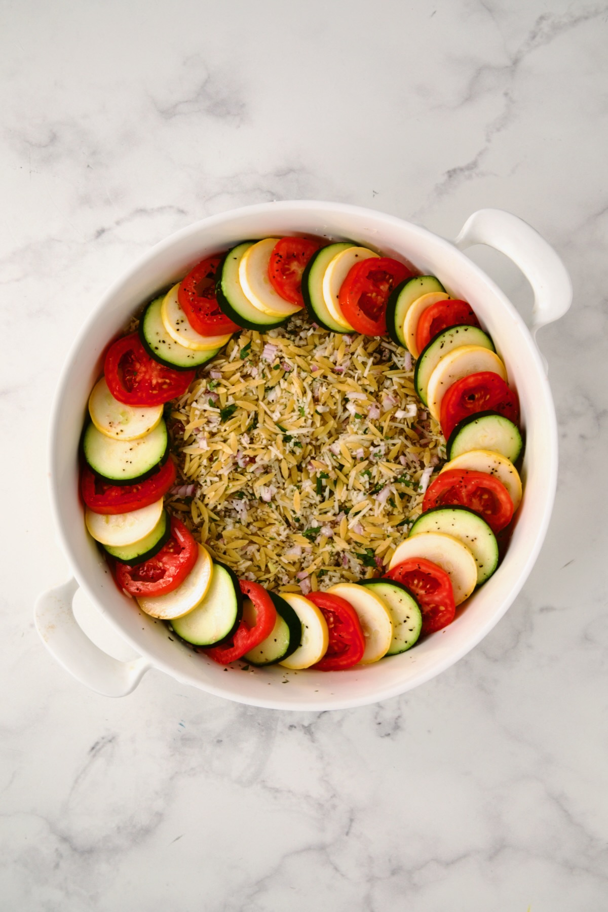 Ingredients for tian in round white baking dish with one circle of yellow squash, zucchini and tomatoes.