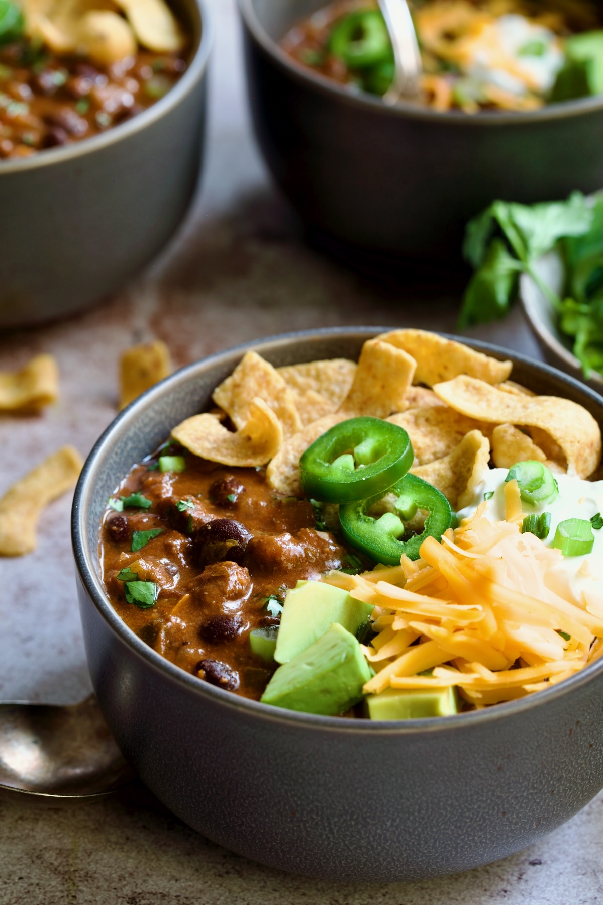 One serving of Beef and Chorizo Chili in gray bowl garnished with avocado, cheddar cheese, sour cream, jalapeno slices and corn chips.
