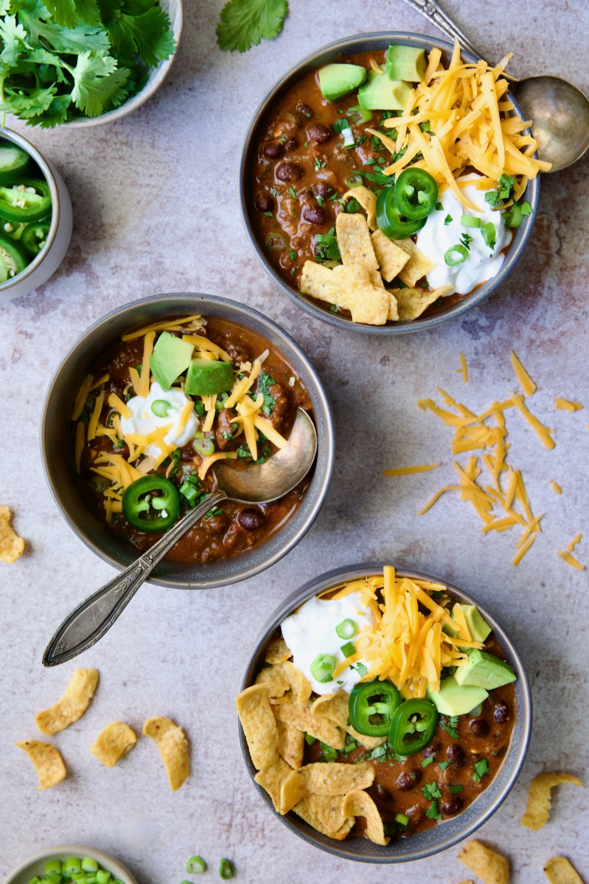 Three servings of Beef and Chorizo Chili in gray bowl garnished with avocado, cheddar cheese, sour cream, jalapeno slices and corn chips.