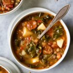 One serving of Spanish Lentil Potato Soup in white bowl with soup spoon with lentils scattered about.