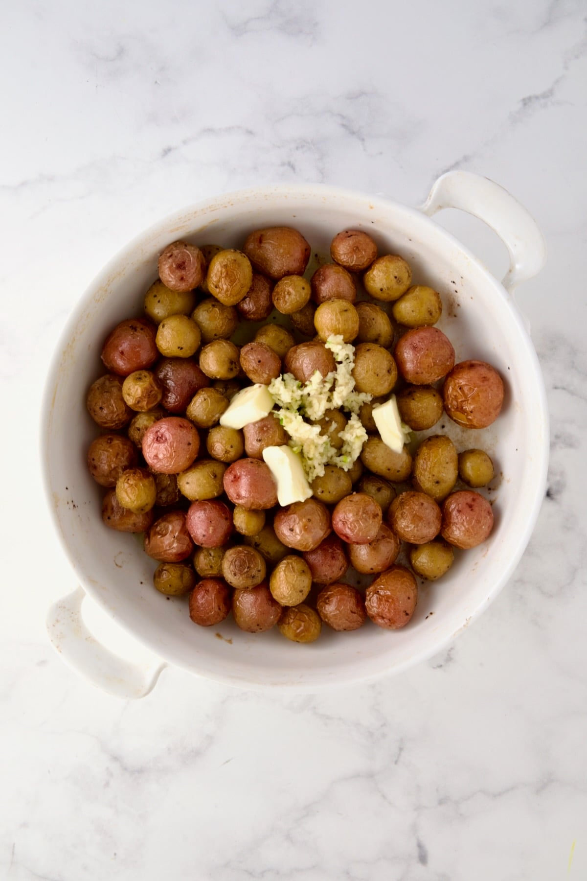 Butter and garlic added to roasted mini potatoes in round white baking dish.