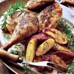 Roast Chicken Half with Fingerling Potatoes on gray-rimmed platter with. meat fork garnished with fresh rosemary and thyme.