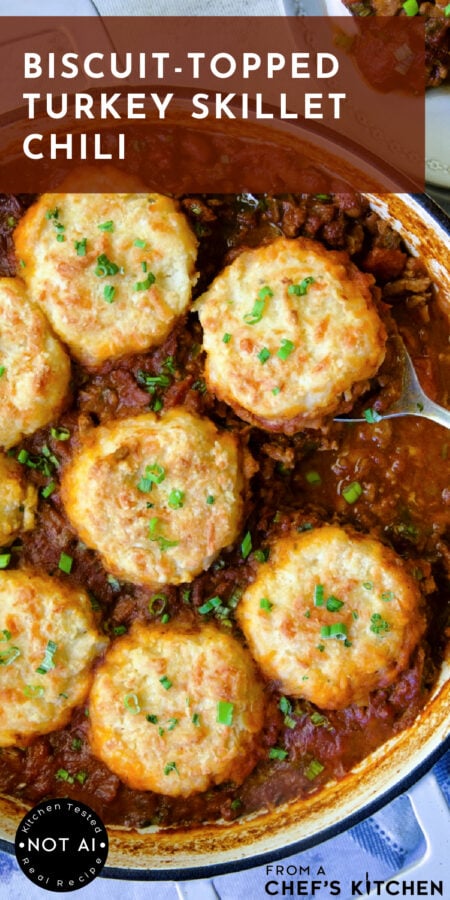 Pinterest graphic for Biscuit-Topped Turkey Skillet Chili showing it in a white enamel cast iron skillet with serving spoon.