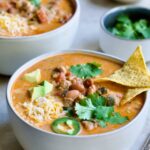 One serving of Creamy Taco Soup in white bowl garnished with tortilla chips, avocado, cheese and jalapeno slices.