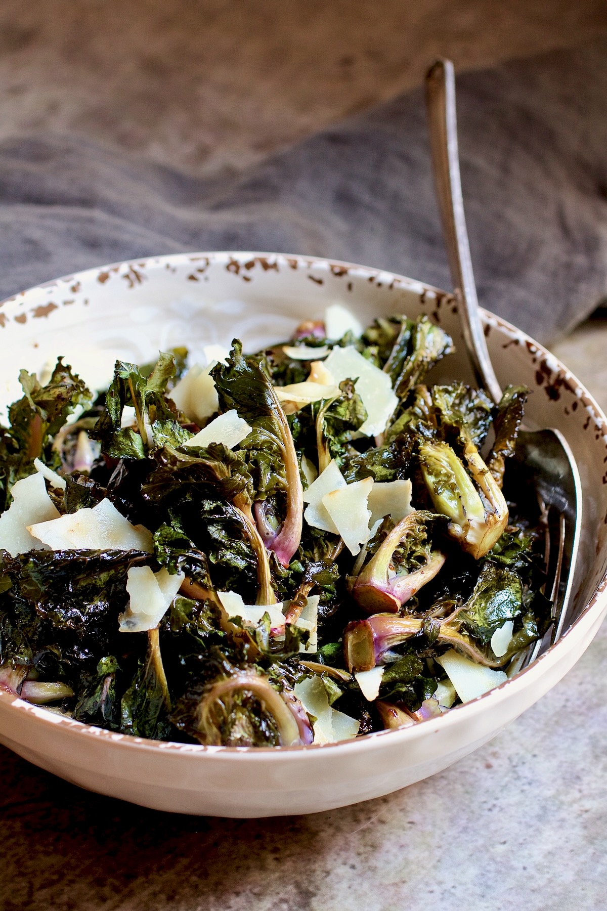 Roasted kale sprouts with shaved Parmesan cheese in white bowl with serving fork.