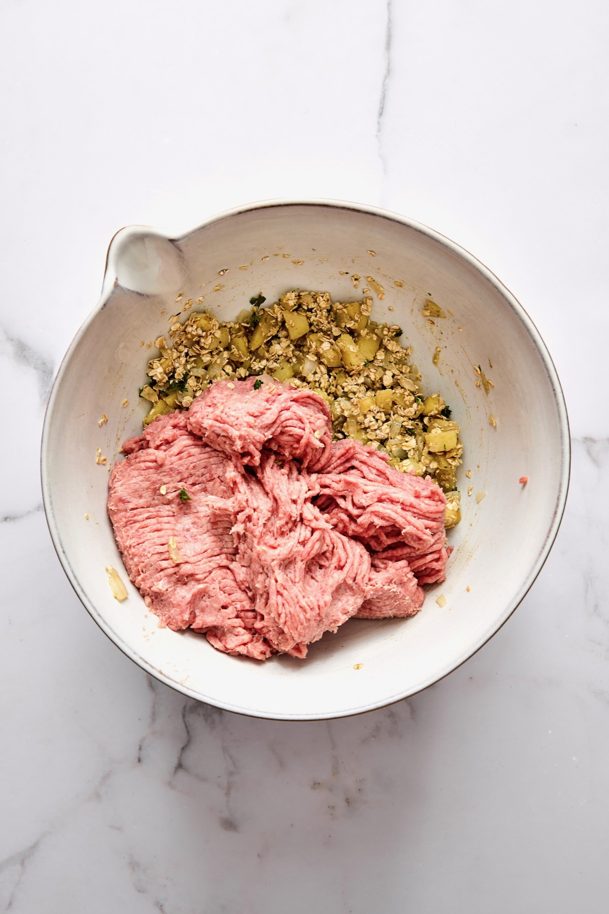 Ground turkey added to cooked onions, apples, oatmeal, parsley and egg in white mixing bowl.