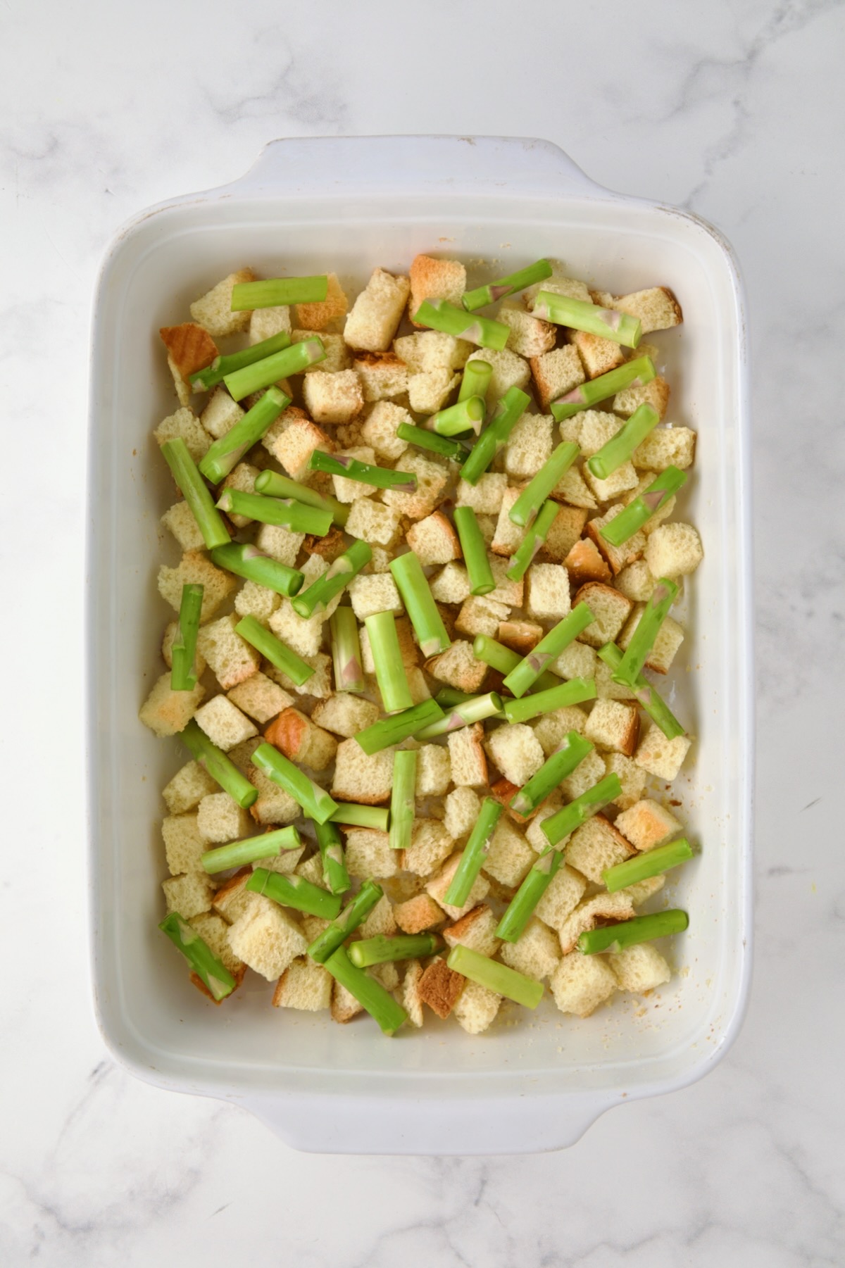 Half of the asparagus pieces over the brioche bread cubes in white rectangular baking dish.