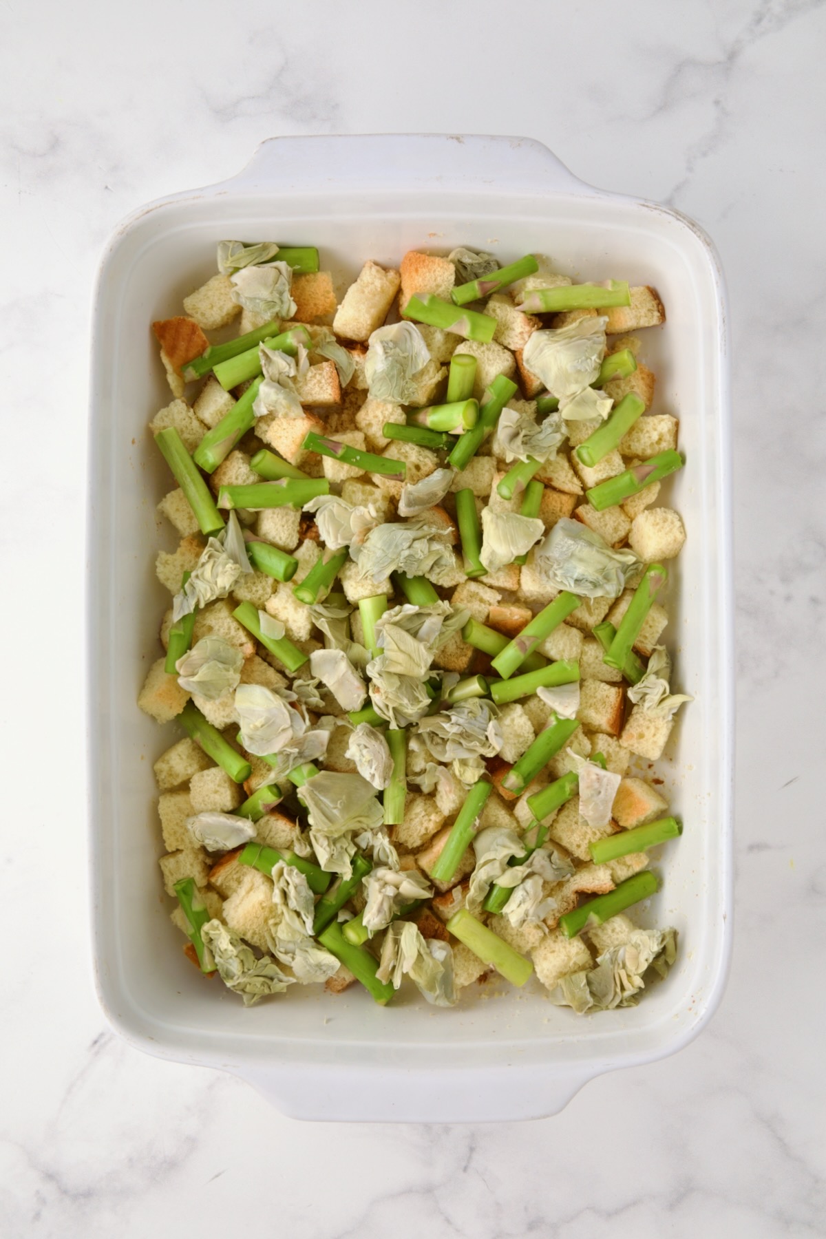 Half of the artichoke hearts sprinkled over the asparagus and brioche cubes in white rectangular baking dish.