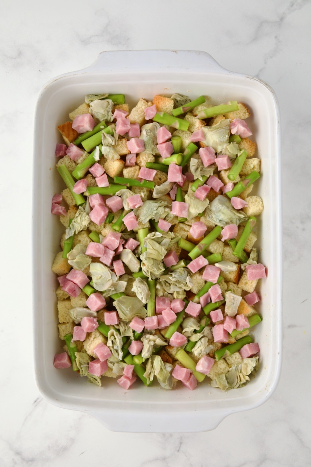 Half of the ham sprinkled over the artichoke hearts, asparagus and bread cubes in white rectangular baking dish.