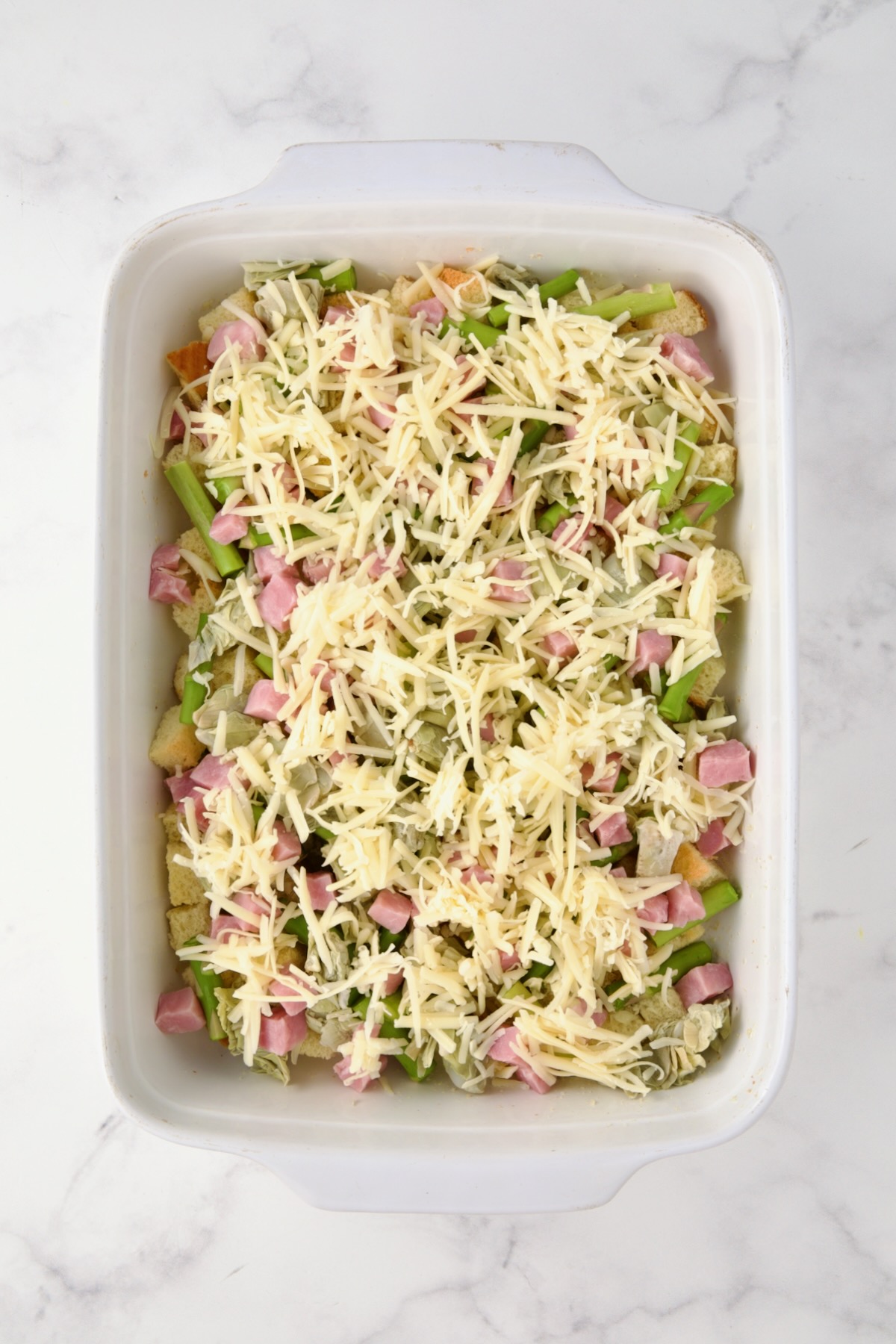 Half of the cheese sprinkled over the ham, artichoke hearts, asparagus and brioche cubes in white rectangular baking dish.