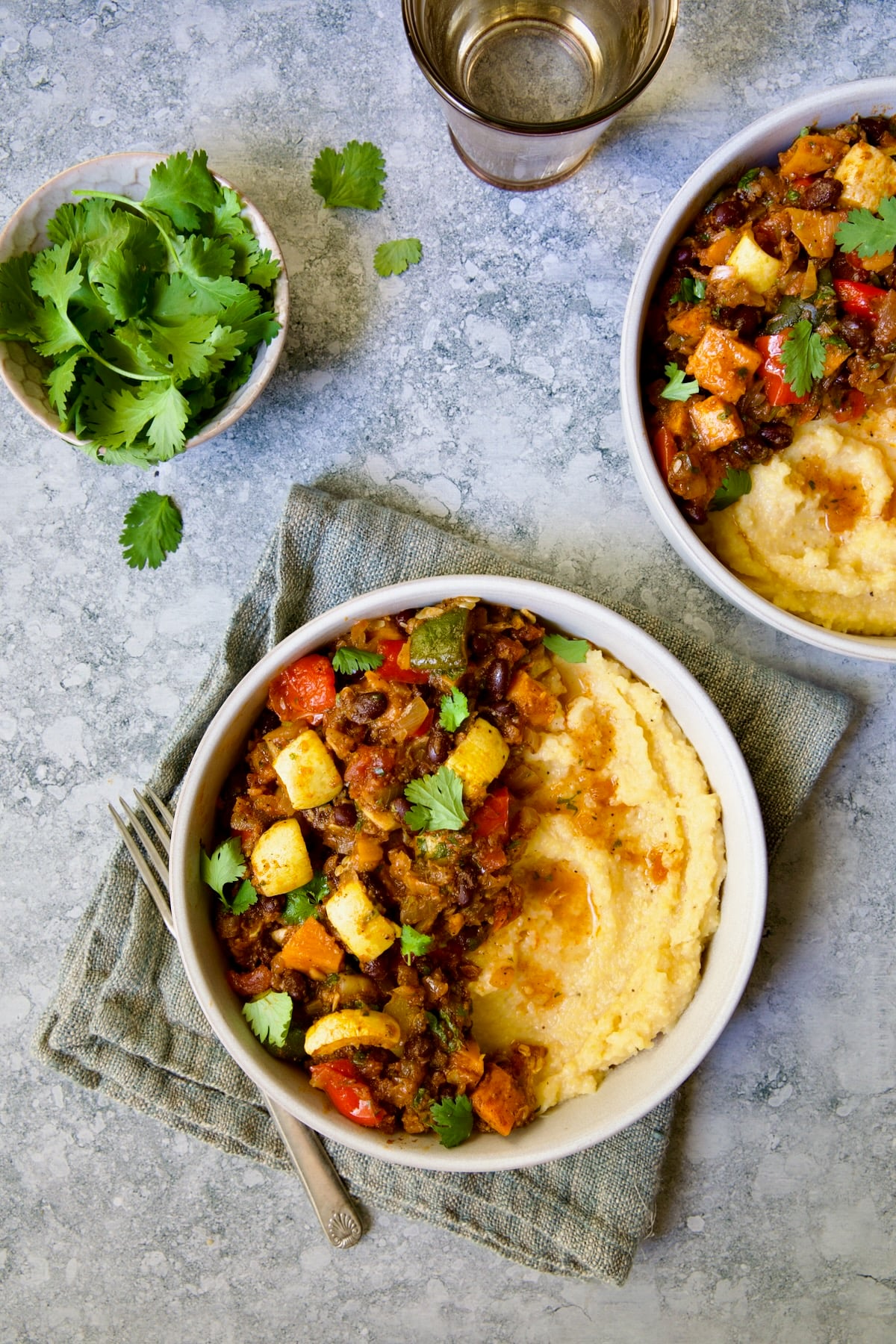 Two servings of Roasted Vegetable Stew with Polenta in white bowls on blue napkin garnished with fresh cilantro leaves.