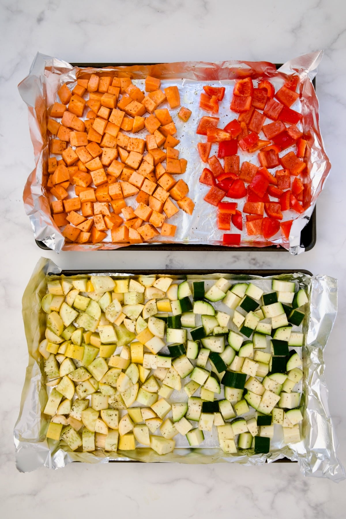Cubed sweet potato, red bell pepper, yellow squash and zucchini on aluminum foil-lined rimmed sheet pans before being roasted.