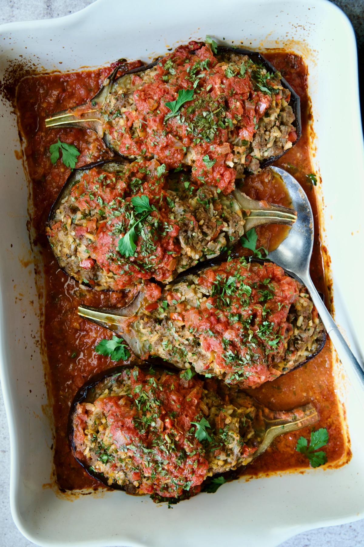 Four baked Stuffed Eggplant halves set a top tomato sauce in white rectangular baking dish with serving spoon.
