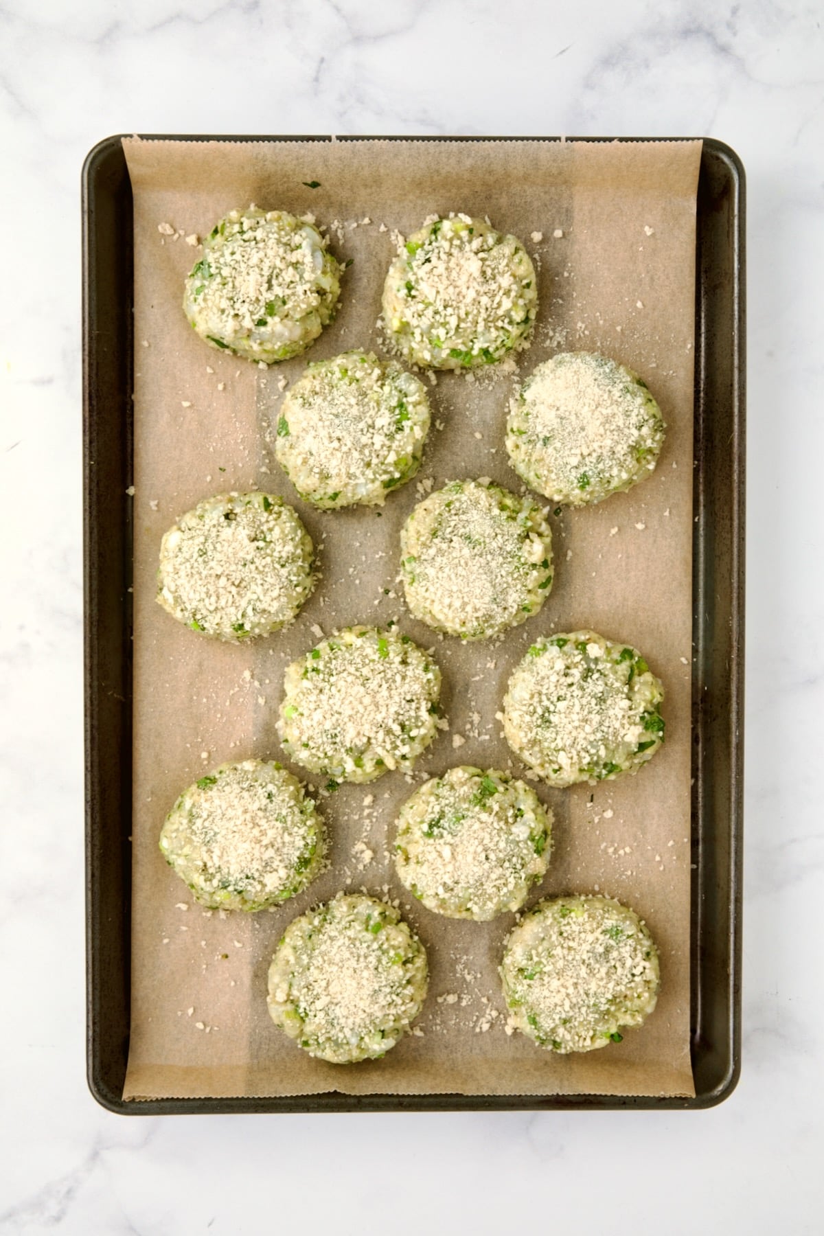 12 uncooked shrimp cakes on parchment paper lined rimmed baking sheet with panko sprinkled over.