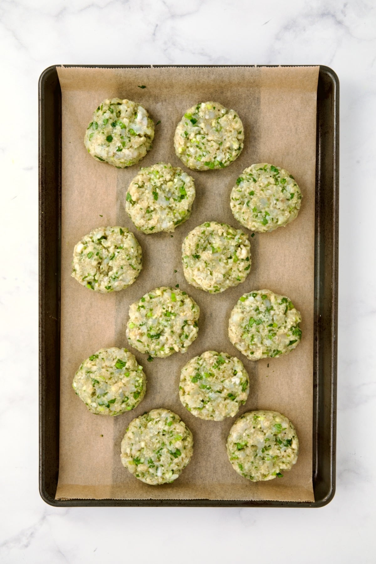 12 uncooked shrimp cakes on parchment paper lined rimmed baking sheet.