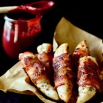 Bacon-Wrapped Chicken Tenders in brown paper lined basket with Raspberry Chipotle Sauce in the background.